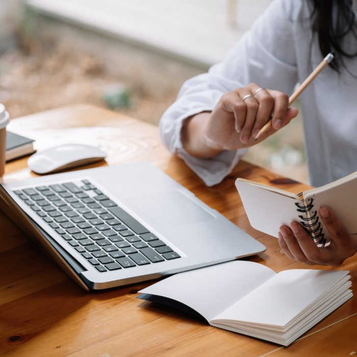 A person sits at a wooden table, holding a spiral-bound notebook and a pencil, looking intently at the open notebook.

The scene is composed of a close-up view of a person's hands holding a notebook and a pencil. The person appears to be focused on the task of writing or reading.  The notebook is positioned centrally, with hands holding it open, and a computer laptop lies on the table beside it. The lighting is soft and ambient, highlighting the person's hands and the notebook. The wooden table serves as a neutral backdrop, and the focus is squarely on the person's actions and the notebook.

The primary subject is a person, likely an adult, whose hands and upper body are visible. The person is holding a spiral-bound notebook and a pencil, appearing to be deeply engaged in reading or writing.  The person's attire is a light-colored, likely white or beige, long-sleeved shirt, and the hands and upper arm portion of the person are visible.  No other details of the person are visible beyond the hands and upper torso.

The artistic medium is photography. The technical execution is skillful, with the image focusing on a soft focus on the notebook, and hands holding it. The style is straightforward, naturalistic, and simple, capturing a casual moment of focus.  The creative choices emphasize the quiet focus of the person engaged in their work. The focus is on the moment and simple details, without artistic embellishments or special effects.

The setting is a simple, wooden table, likely indoors. The lighting conditions are soft and natural, casting no harsh shadows. The environment appears to be a workspace or home office, providing a sense of calmness and productivity. The wooden surface and soft lighting contribute to the overall atmosphere. The setting is uncluttered, allowing the focus to remain on the person and the activity.