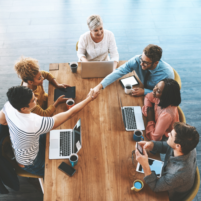 A high-angle shot captures a group of diverse people seated around a wooden table, engaged in a business meeting or collaboration session, with a handshake taking place in the center.

The scene composition is a top-down view of a rectangular table where several people are seated, creating a circular arrangement around the table.  Hands are outstretched in a handshake, signifying a collaborative or agreement-making moment. The table is filled with laptops, coffee cups, and other digital devices, suggesting a workplace setting. The positioning of the individuals around the table creates a sense of focused interaction and collaboration. The framing is tight, centering the action of the handshake, highlighting the group's interaction and activity.

The key subjects are various adults, comprising a diverse group in terms of gender and ethnicity. Their attire suggests a professional or business context. Facial expressions are varied but generally show engagement and attention. One particular interaction of the people is the handshake, and the overall mood seems collaborative and positive.

The artistic medium is photography, capturing a candid moment in a contemporary office setting.  The technical execution is clear, showing sharp focus on the subjects and the table, showcasing a professional quality image. The style is documentary in nature, showcasing a realistic depiction of a business interaction. The creative choice is an overhead shot providing a comprehensive view of the group and interaction at the table, offering an insight into the workplace dynamic.

The setting is an office environment.  The lighting is bright and evenly distributed, creating a clean and neutral atmosphere. The table and its elements are well-lit, and the neutral color tones of the room complement the overall presentation of the image. The atmosphere is one of professional and collaborative engagement, suited for a work setting.