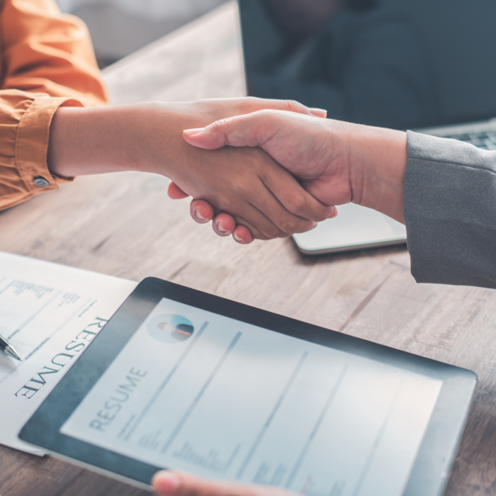 Two people shake hands across a desk, signalling the successful conclusion of a job interview or meeting. On the table are a printed CV, a tablet displaying a digital version of a resume, and an open laptop, reflecting a modern, hybrid approach to recruitment. One person is wearing an orange shirt, while the other is in a grey blazer, suggesting a formal but approachable setting. The close-up focus on the handshake captures a moment of agreement or a potential job offer, emphasising professionalism, trust, and mutual respect.