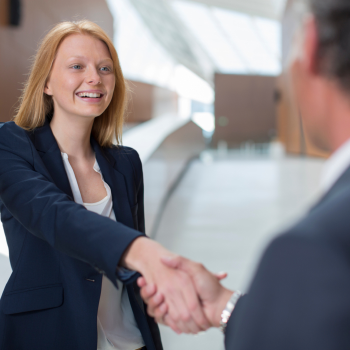 A young woman with long, straight blonde hair, dressed in a navy blazer and white blouse, extends her hand in a handshake while smiling warmly. She is standing in a modern, well-lit building with high ceilings and large windows that allow natural light to flood the space. Her posture is confident and welcoming. In the foreground, a man in a dark suit, seen from behind, is returning the handshake. The setting appears professional, with clean architectural lines and a bright, airy atmosphere.