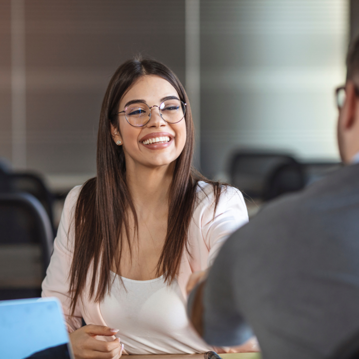 A woman with long brown hair and glasses is seated at a table, smiling and shaking hands with a man. The woman is wearing a light pink or peach-colored blazer over a white top. The man is wearing a dark gray or charcoal-colored business suit. The image appears to be from a business meeting or interview, as both individuals are dressed in professional attire. A laptop is visible on the table in front of the woman. The background is out of focus, showing a portion of an office space. The woman is in the foreground, and the man's upper body, and part of his head are visible.