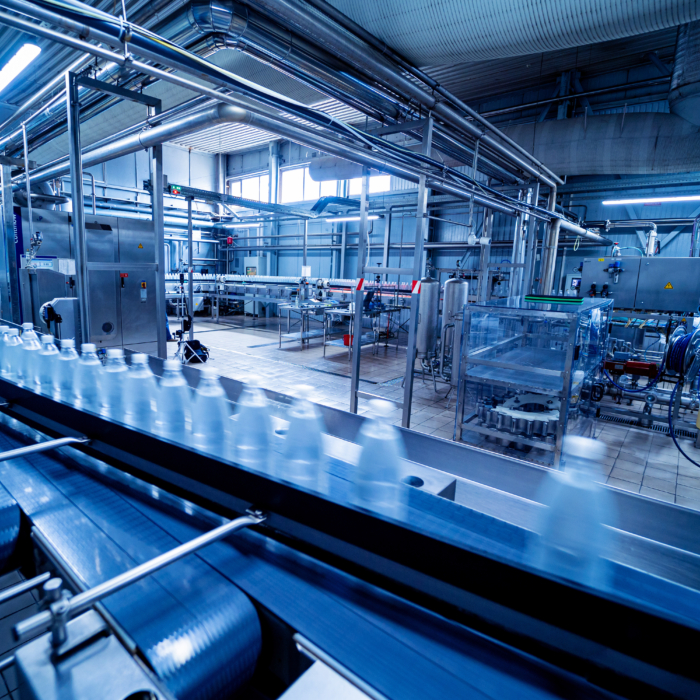 Full indoor shot of a beverage bottling or processing facility. 

A series of conveyors crisscross the floor, transporting clear plastic bottles. The conveyors are made of metal and appear to be in motion. 

The facility is filled with various stainless steel machinery and equipment, including tanks, pumps, and other processing devices. 

Piping and metal supports crisscross the ceiling and walls. 

The lighting in the facility is bright, and the overall color scheme is a cool, blue-toned palette. The floor is tiled. The focus is on the active bottling line.