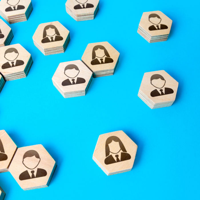 Wooden hexagonal tiles featuring male and female business avatars are arranged on a bright blue background. Some tiles are grouped together while others are separated, symbolising workforce segmentation, recruitment, or staffing flexibility. The visual conveys concepts related to human resources, talent selection, and strategic team placement.