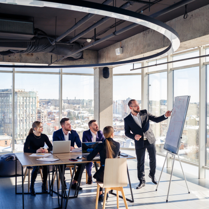 A group of business professionals are gathered in a modern, bright office space for a presentation.
A man in a dark suit is standing at a whiteboard, pointing to it, leading a presentation to a group of colleagues. The group is seated around a light-colored wooden table, and several individuals are seated at the table with laptops open while others are standing.
The room features large windows overlooking a cityscape, and a large, circular ceiling fixture hangs above. The space is brightly lit and airy, suggesting a modern office environment. The layout is designed for efficient meetings and presentations.