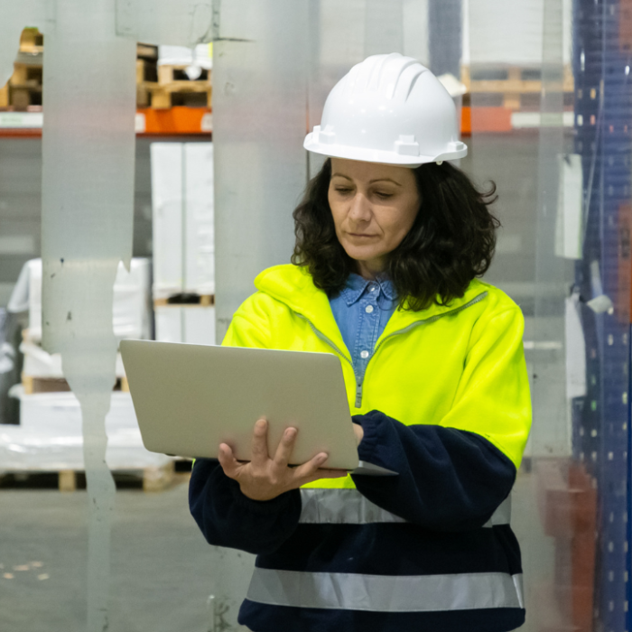 A woman stands in a bustling warehouse environment, dressed in a high-visibility yellow safety jacket over a dark navy blue sweatshirt, which contrasts sharply against the bright hue of her outer layer. Her jacket features reflective stripes that emphasize safety protocols. The woman has medium-length, naturally curly dark brown hair, framing her face gently, and is focused intently on a silver laptop she holds in both hands. 

She wears a white hard hat, typical for warehouse settings to ensure safety, which adds a layer of professionalism to her demeanor. The environment around her is structured and organized, with large wooden pallets stacked in the background. Various items are visible on the shelves, suggesting a diverse inventory. Clear plastic sheeting hangs loosely from ceiling to floor, possibly indicating areas that are being maintained or sectioned off.

The flooring is concrete, which reflects the industrial nature of the workspace, while the overall lighting is bright, enhancing visibility for any tasks being performed. Small details like the textures of the materials—the smoothness of the laptop, the weave of the fabric in her jacket, and the roughness of the concrete—add depth to the scene. Her expression conveys concentration and responsibility, hinting at the importance of the task at hand in this active warehouse setting.