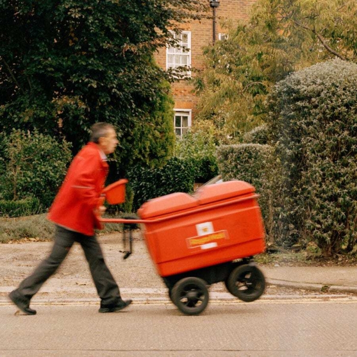A Royal Mail postal worker in a high-visibility jacket walks along a leafy street pushing a red mail trolley while delivering letters.