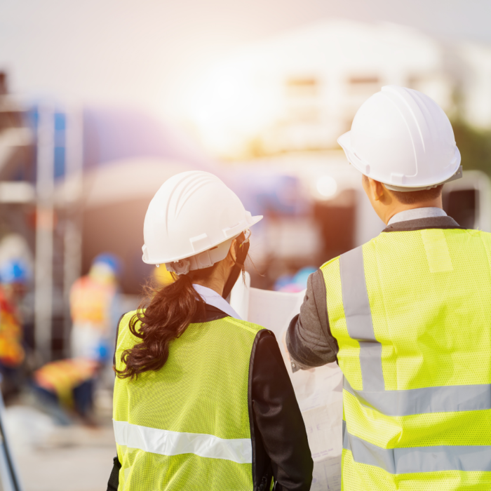Two engineers wearing hard hats and reflective vests review plans on a construction site. The scene highlights teamwork, project management, and safety in the building and infrastructure industry.