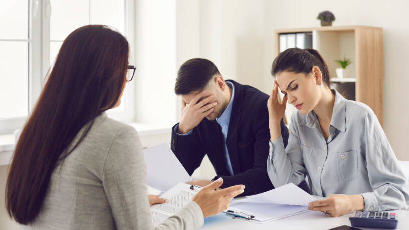 A woman and a man, both appearing distressed, sit at a table with a third person, likely a counsellor or advisor. 

The woman and man both have their heads in their hands or near their foreheads, suggesting distress or frustration.  Documents and a calculator are on the table. 

The woman and man are wearing business-casual attire, consisting of business shirts and pants.  The third person, partially visible, is wearing a light grey top.  

The setting appears to be an office or consulting room, with a window visible in the background. The overall impression is of a tense or difficult conversation regarding problems.