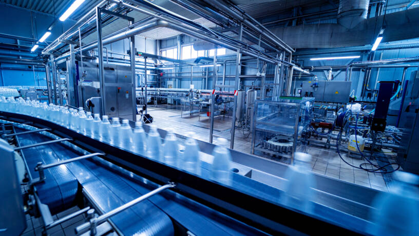 Full indoor shot of a beverage bottling or processing facility. 

A series of conveyors crisscross the floor, transporting clear plastic bottles. The conveyors are made of metal and appear to be in motion. 

The facility is filled with various stainless steel machinery and equipment, including tanks, pumps, and other processing devices. 

Piping and metal supports crisscross the ceiling and walls. 

The lighting in the facility is bright, and the overall color scheme is a cool, blue-toned palette. The floor is tiled. The focus is on the active bottling line.