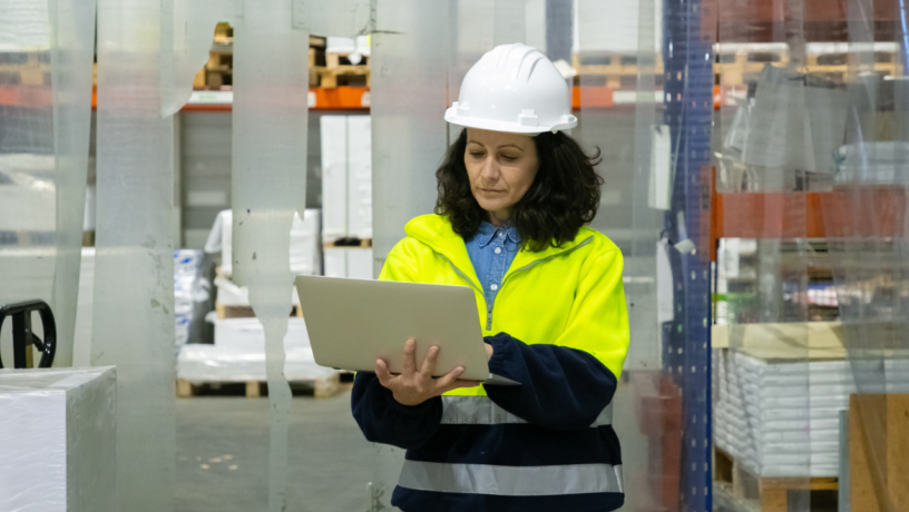 A woman stands in a bustling warehouse environment, dressed in a high-visibility yellow safety jacket over a dark navy blue sweatshirt, which contrasts sharply against the bright hue of her outer layer. Her jacket features reflective stripes that emphasize safety protocols. The woman has medium-length, naturally curly dark brown hair, framing her face gently, and is focused intently on a silver laptop she holds in both hands. 

She wears a white hard hat, typical for warehouse settings to ensure safety, which adds a layer of professionalism to her demeanor. The environment around her is structured and organized, with large wooden pallets stacked in the background. Various items are visible on the shelves, suggesting a diverse inventory. Clear plastic sheeting hangs loosely from ceiling to floor, possibly indicating areas that are being maintained or sectioned off.

The flooring is concrete, which reflects the industrial nature of the workspace, while the overall lighting is bright, enhancing visibility for any tasks being performed. Small details like the textures of the materials—the smoothness of the laptop, the weave of the fabric in her jacket, and the roughness of the concrete—add depth to the scene. Her expression conveys concentration and responsibility, hinting at the importance of the task at hand in this active warehouse setting.